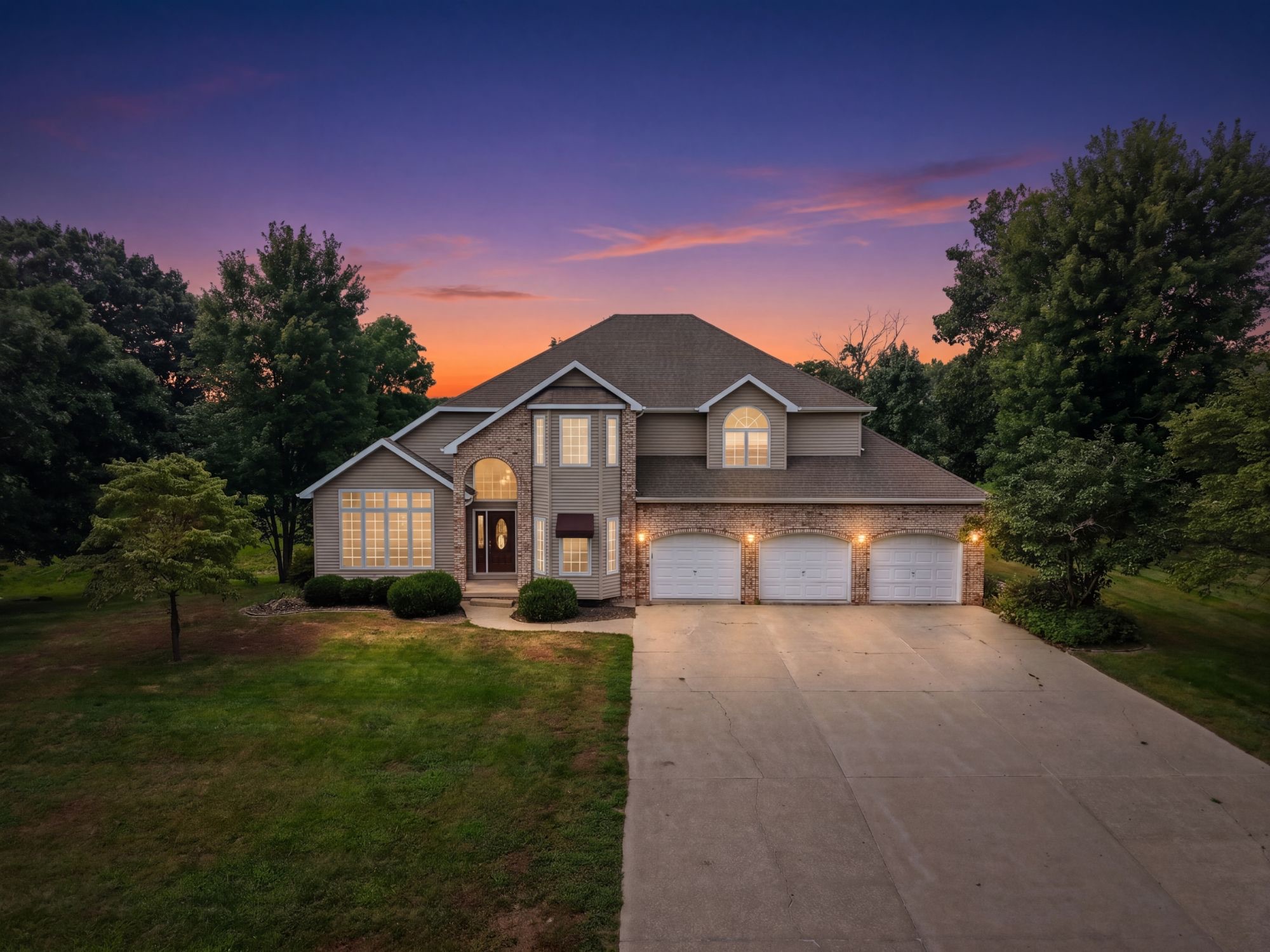 Two-story brick home with a three-car garage at twilight, windows and porch lights glowing